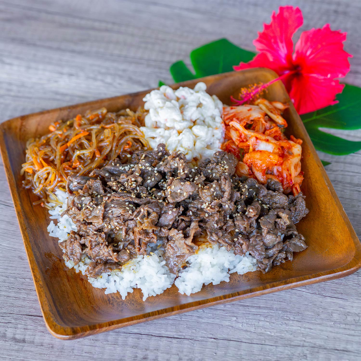 A square wooden plate sits on a light wood surface, filled with a generous portion of thinly sliced bulgogi beef sprinkled with sesame seeds, all served over a bed of white rice. The plate also holds a serving of translucent glass noodles with shredded carrots, a scoop of creamy macaroni salad, and a side of spicy red kimchi. Decorative green tropical leaves and a pink hibiscus flower frame the upper corner of the plate.