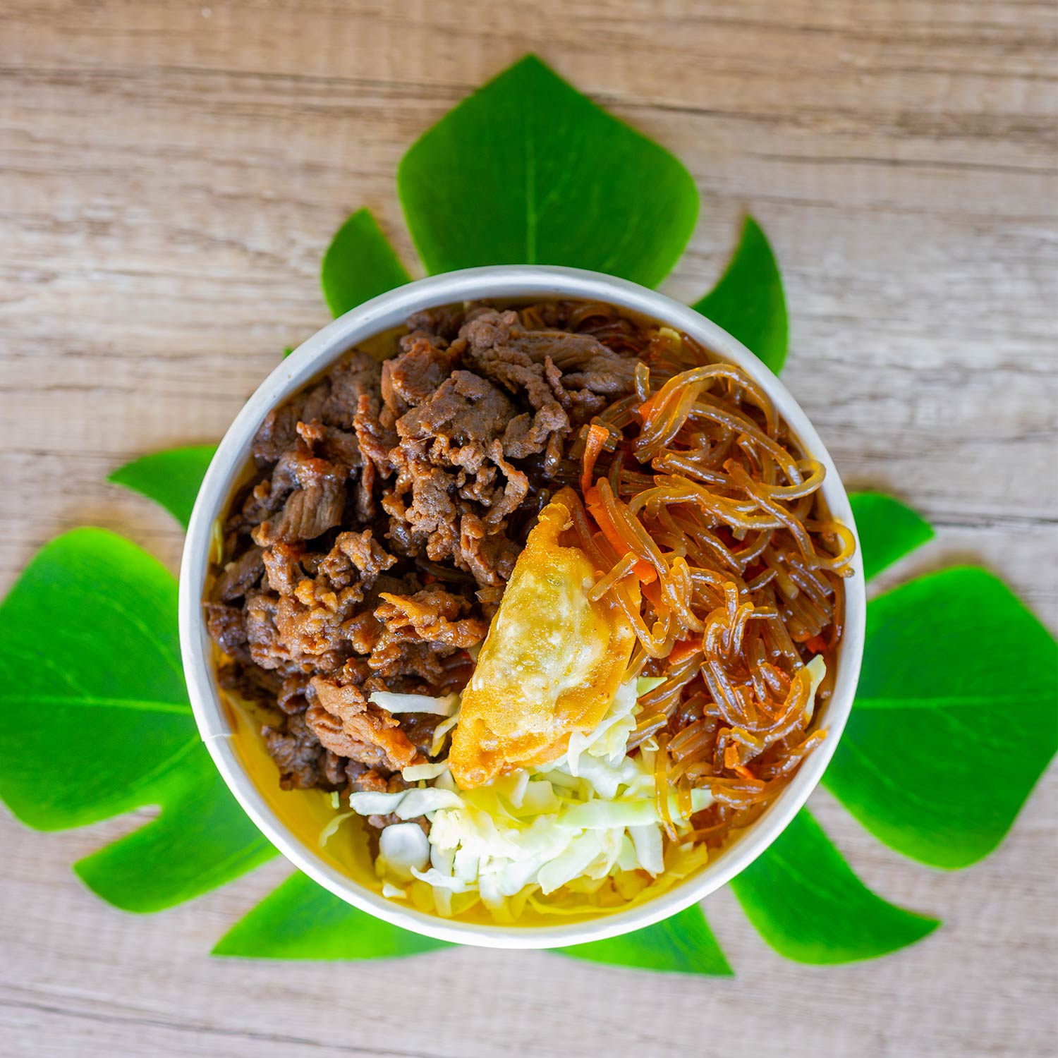 A round bowl is filled with glass noodles and shredded cabbage, topped with thinly sliced beef bulgogi and a single golden fried dumpling. The bowl is arranged on a wooden tabletop, with several green leaf cutouts placed underneath to form a decorative floral-like background.
