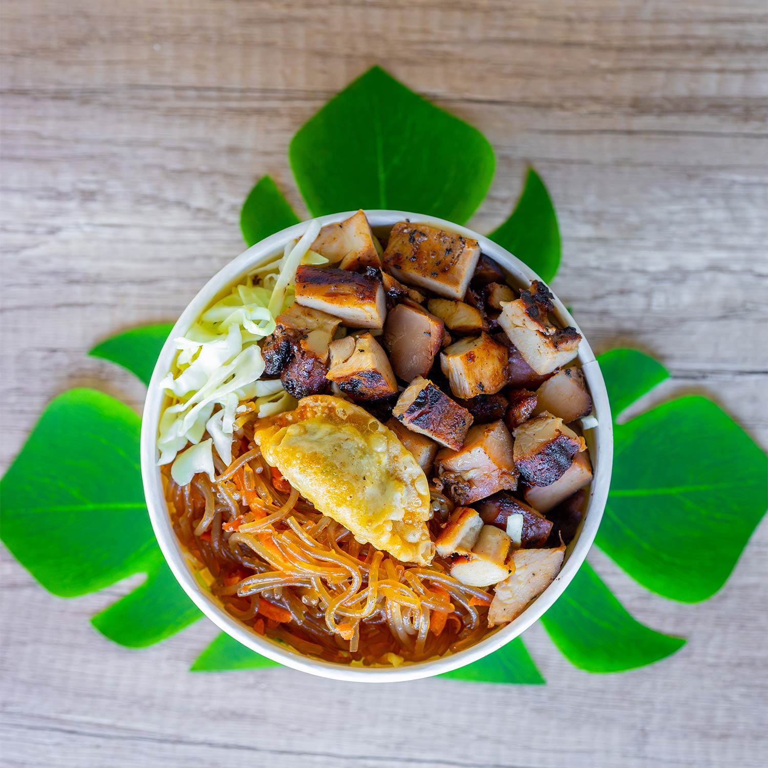 A round bowl on a wooden surface contains grilled, cubed pieces of boneless chicken, translucent glass noodles with carrots, shredded cabbage, and a single fried dumpling. The bowl is positioned above several green leaf cutouts arranged in a circular pattern, giving a floral appearance to the background.