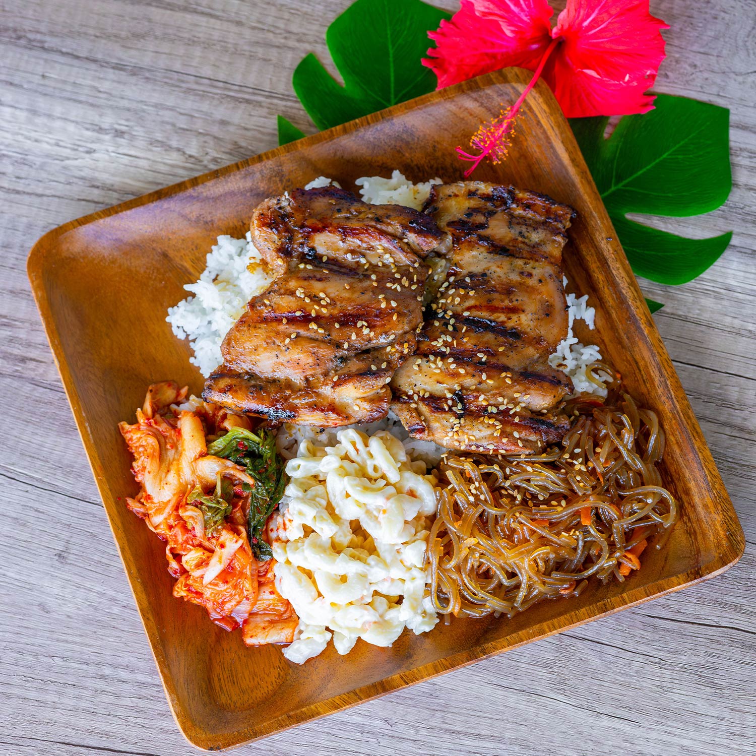 This image features a wooden square plate containing grilled boneless chicken thighs topped with sesame seeds, accompanied by a scoop of white rice, creamy macaroni salad, kimchi, and a portion of clear glass noodles with sesame seeds. Green tropical leaves and a pink hibiscus flower are used as garnishes next to the plate, which rests on a pale wooden surface.
