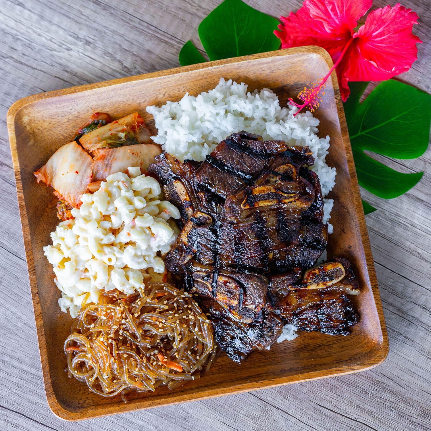 A square wooden plate is arranged with several foods, including grilled kalbi beef short ribs with a caramelized exterior, a scoop of fluffy white rice, a serving of creamy macaroni salad, kimchi, and a small portion of clear glass noodles sprinkled with sesame seeds. Decorative green leaves and a vibrant pink hibiscus flower are placed beside the plate, all set on a wooden table.