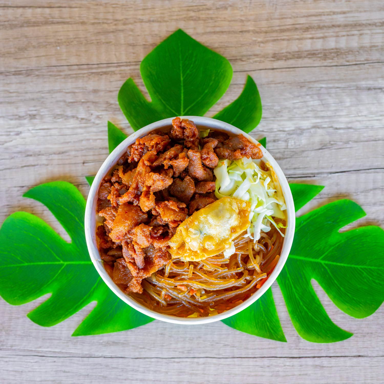 A round bowl filled with crispy pieces of fried pork, finely shredded cabbage, and clear glass noodles sits at the center of the image on a wooden surface. The bowl is also topped with a single golden-brown fried dumpling. Underneath the bowl are several large, bright green tropical leaves arranged in a star pattern for decorative effect.