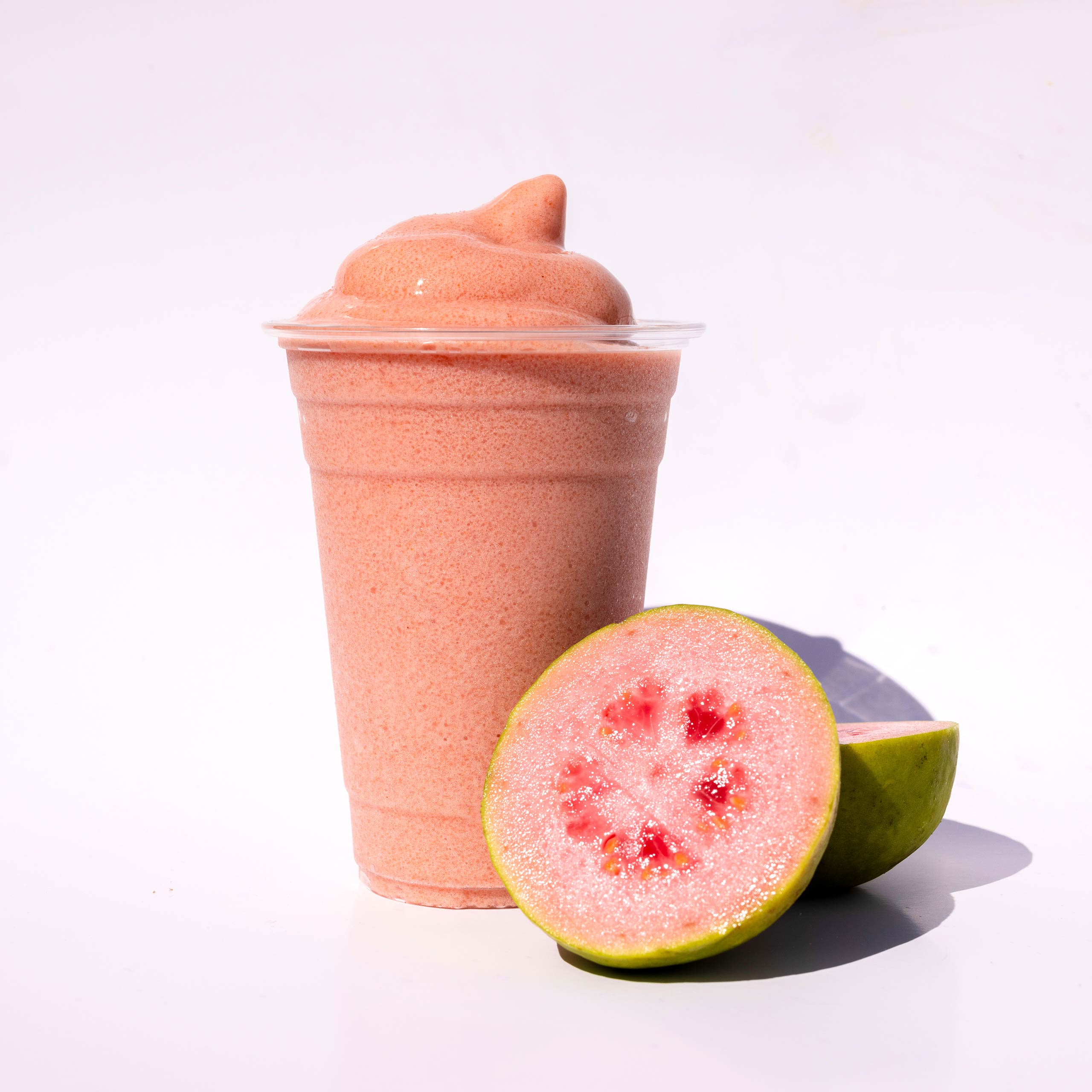 A pink guava slushie in a clear cup, shown next to a fresh guava that has been cut in half.